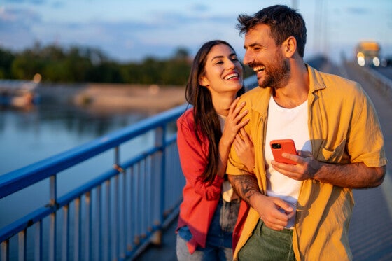 A joyful couple is captured laughing together while sharing a moment on a bridge at sunset, engaging with a phone. Their casual attire reflects a relaxed vibe.