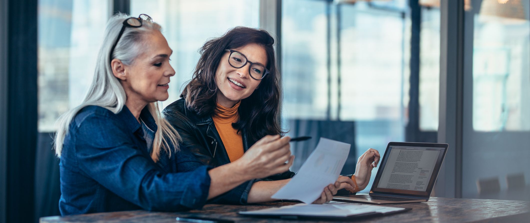 Two women analyzing documents while sitting on a table in office. Woman executives at work in office discussing some paperwork.