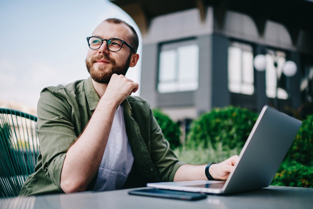 Young handsome male 20 years old in eyeglasses sitting at street cafe during free time with new modern laptop, bearded man freelancer thoughtfully looking aside and thinking about distance work (Young handsome male 20 years old in eyeglasses sitting