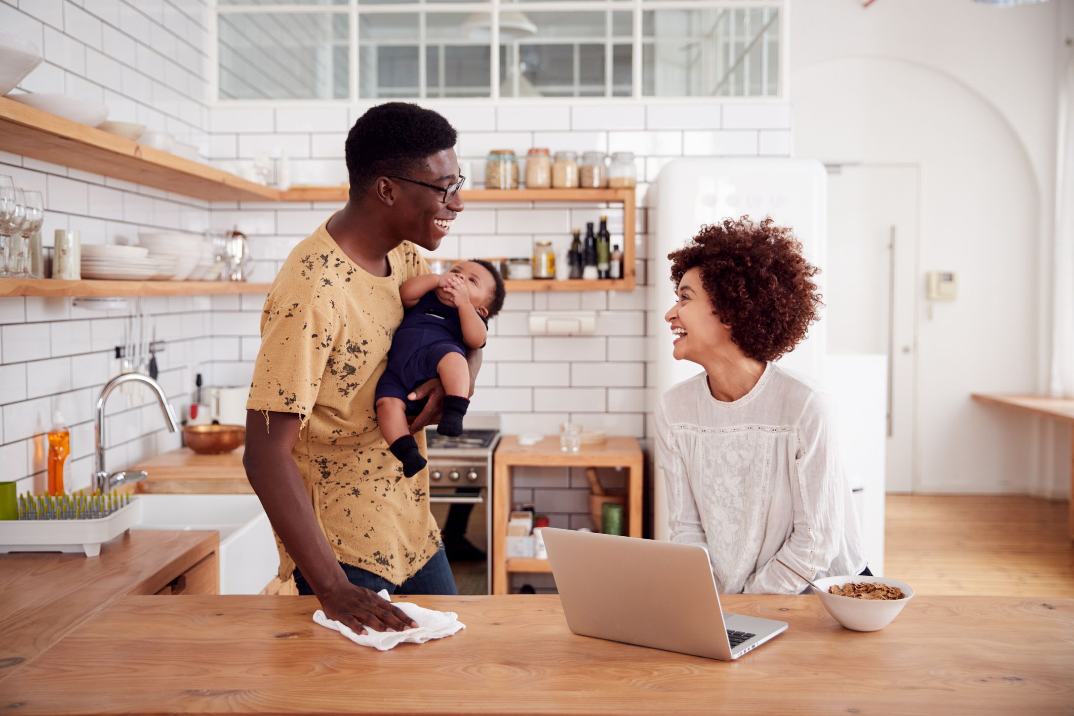 Multi-Tasking Father Holds Baby Son And Cleans Surface As Mother Uses Laptop And Eats Breakfast