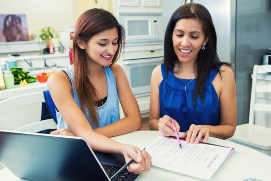 Mother and teenage daughter are in the kitchen researching colleges and filling out university applications.