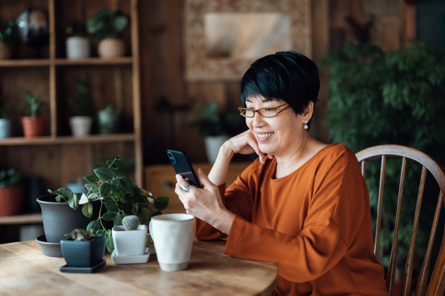 Older Asian woman sitting at kitchen table smiling at cell phone.