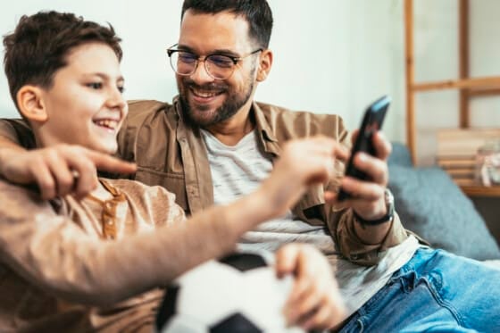 Boy watching soccer match with father at home, sitting on sofa