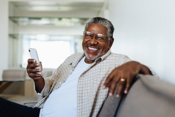 Cheerful man enjoying modern technology at home, sitting comfortably on the couch and smiling joyfully while holding a smartphone