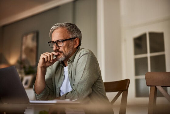 A hardworking mature man freelancer with eyeglasses reading an online project over a laptop, sitting at the wooden table at home.