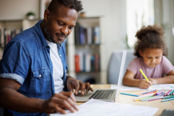 Father working on laptop while his daughter doing homework beside him at home