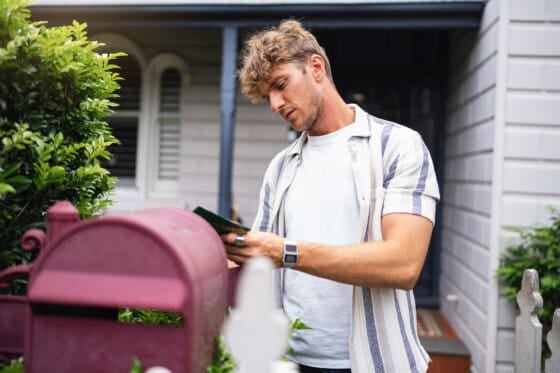 A man wearing casual clothing, standing outside a modern home, checks a mailbox surrounded by greenery. The image conveys daily routines, anticipation, and home residency.