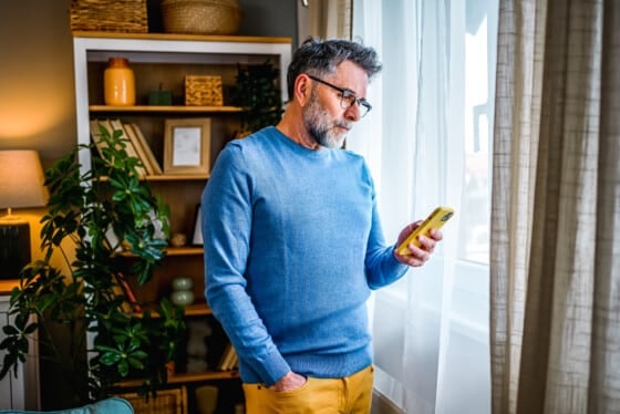 Mature man standing by the window at home and using a mobile phone